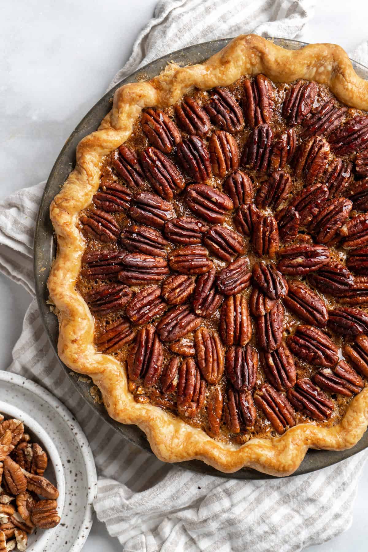 A perfect pecan pie recipe on a white background with a bowl of pecans nearby under a  napkin