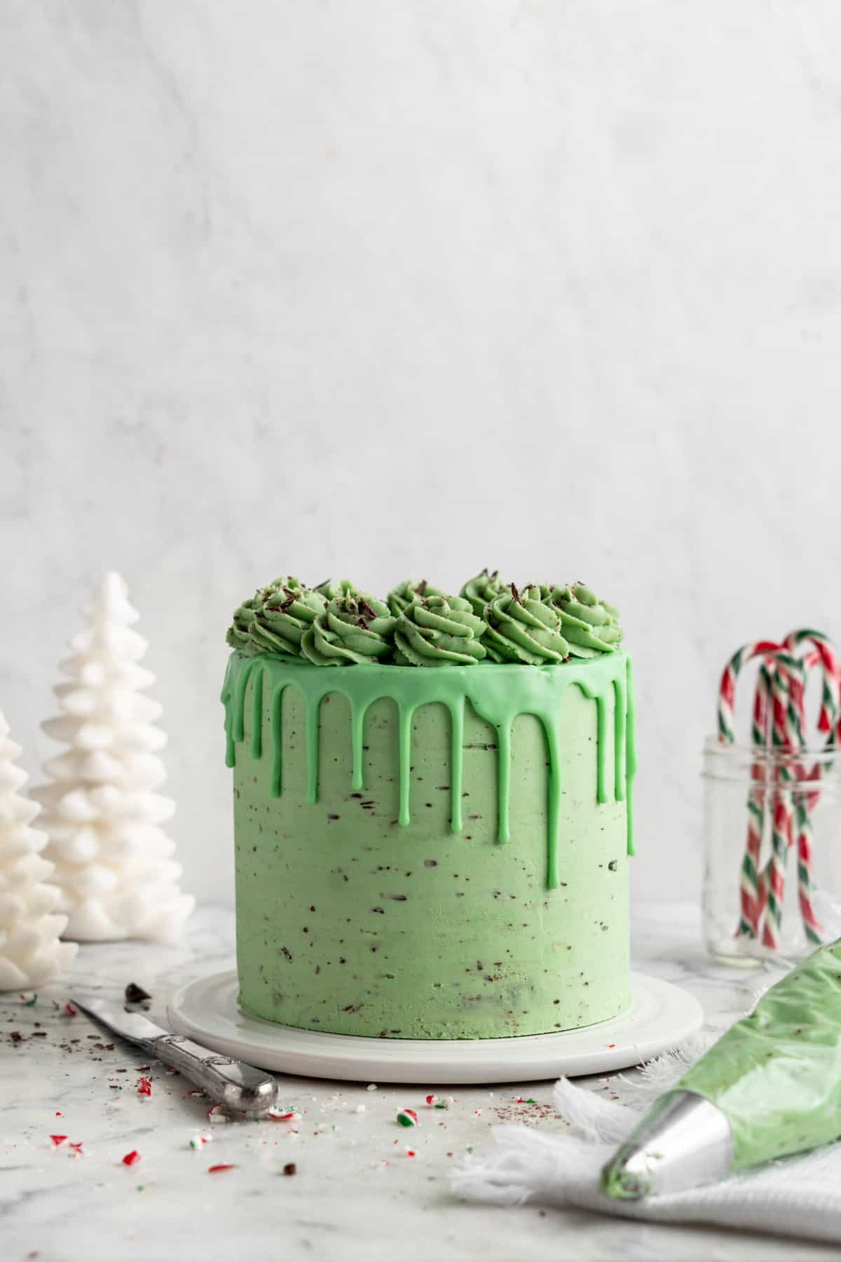 A tall, frosted mint chocolate cake with chocolate specks and a green drip glaze, topped with piped green frosting swirls, styled for Christmas with white trees and candy canes in the background