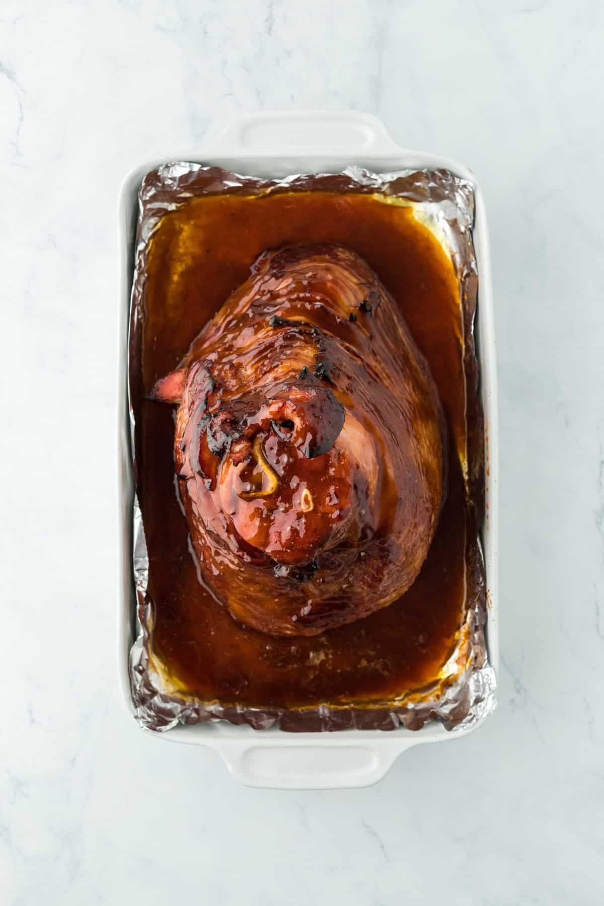 Honey glaze poured over top of a honey baked ham on foil lined baking sheet on white countertop