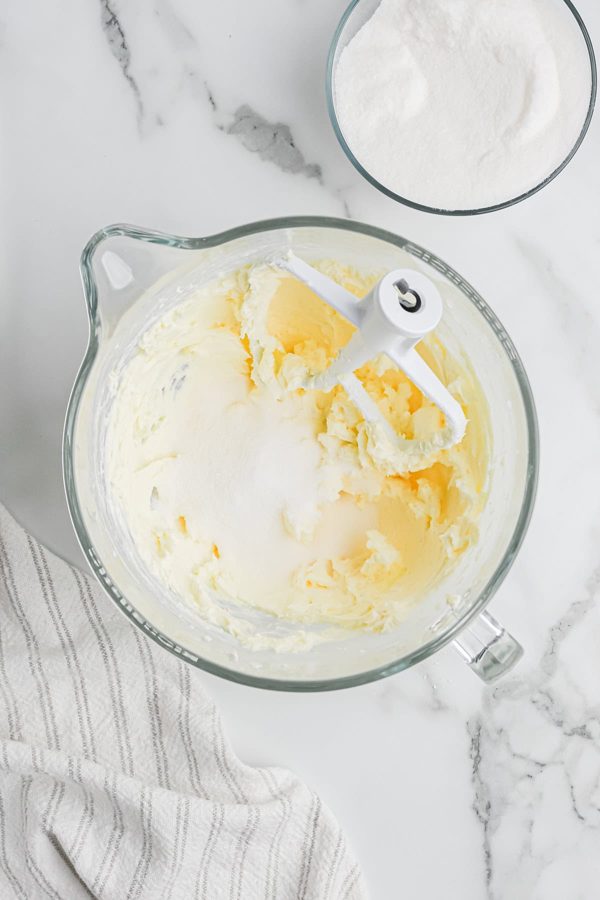 Sugar being added to cream cheese and butter in a clear stand mixer on white background