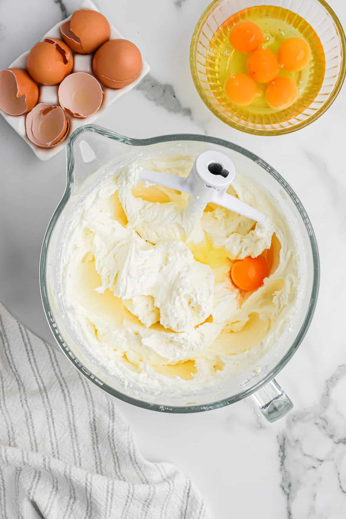 Eggs being added to pound cake batter in clear mixer bowl with eggs nearby on white background