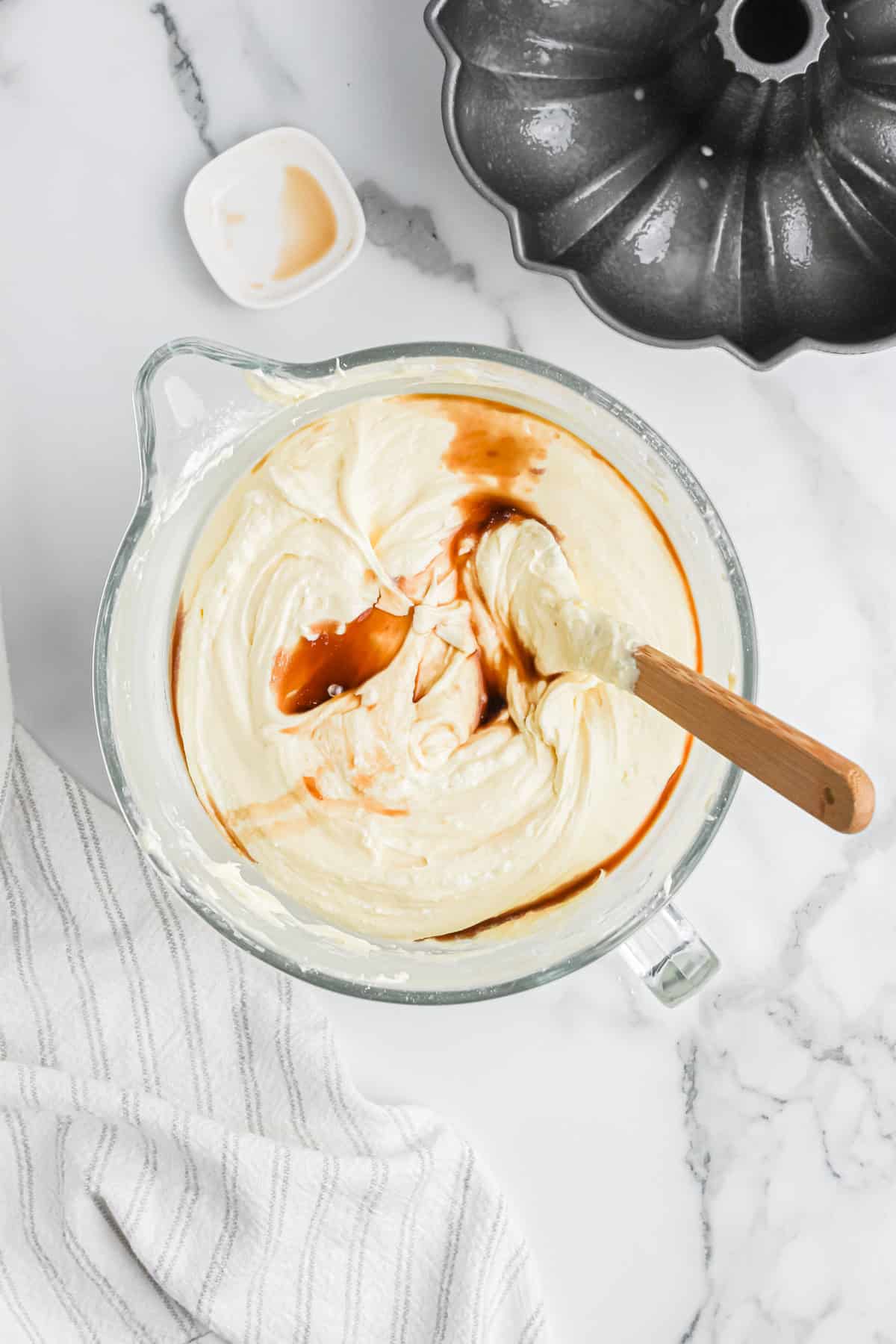 Vanilla extract being added to cream cheese pound cake batter in clear mixer bowl on white background with bundt pan nearby