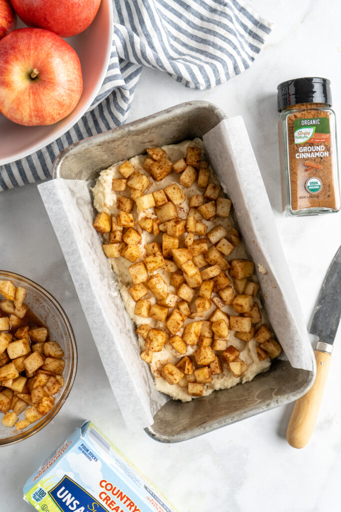 Spiced apples over half bread batter with cinnamon and eggs nearby