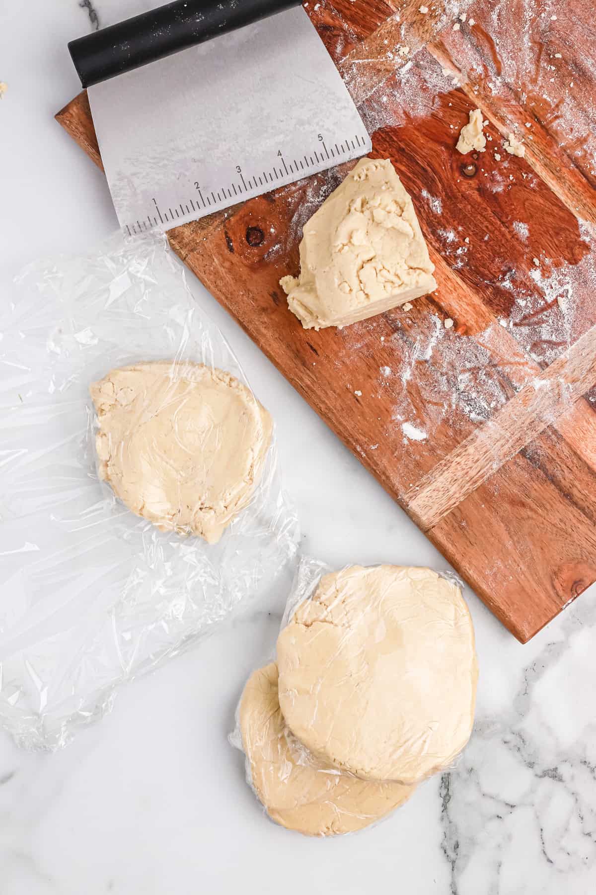 Portions of sugar cookie dough wrapped in plastic wrap on a marble surface next to a floured wooden board and dough scraper