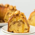 A slice of caramel apple cake on a white plate in front of a whole cake behind on white background