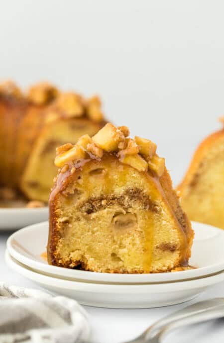 A slice of caramel apple cake on a white plate in front of a whole cake behind on white background