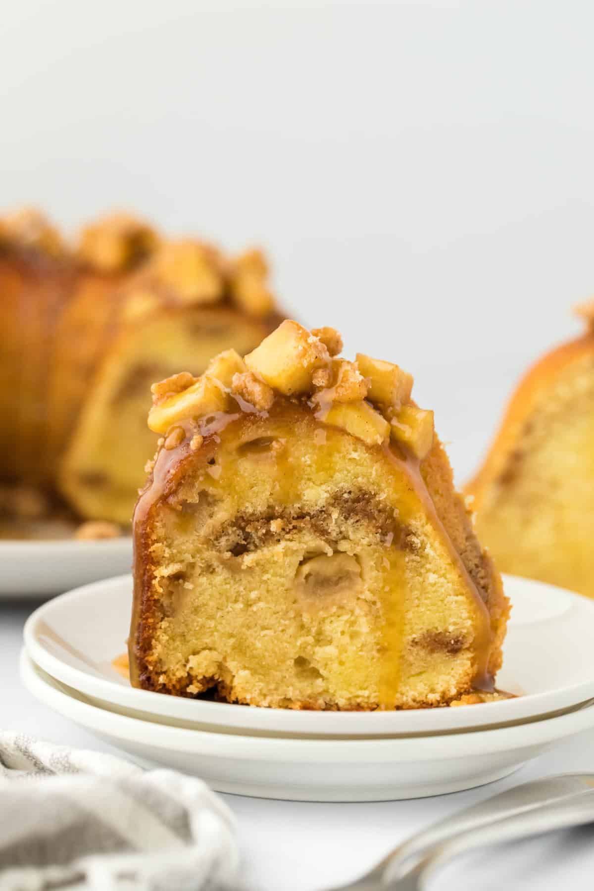 A slice of caramel apple cake on a white plate in front of a whole cake behind on white background