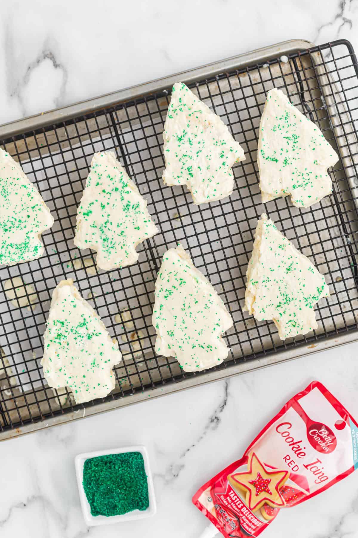 Frosted little debbie christmas tree cakes on a cooling rack sprinkled with green sugar, with red cookie icing visible in the corner