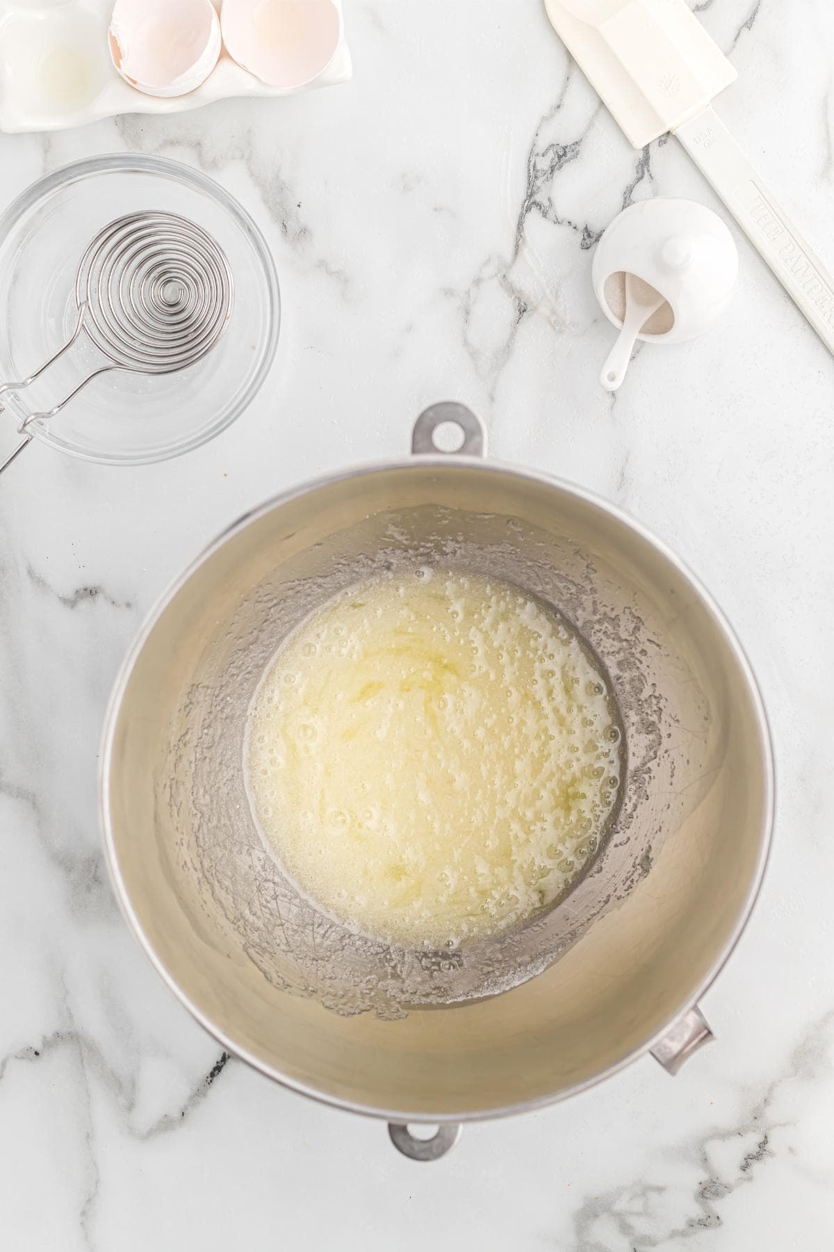 A mixing bowl with bubbly beaten egg whites and sugar sitting on a marble counter surrounded by baking tools