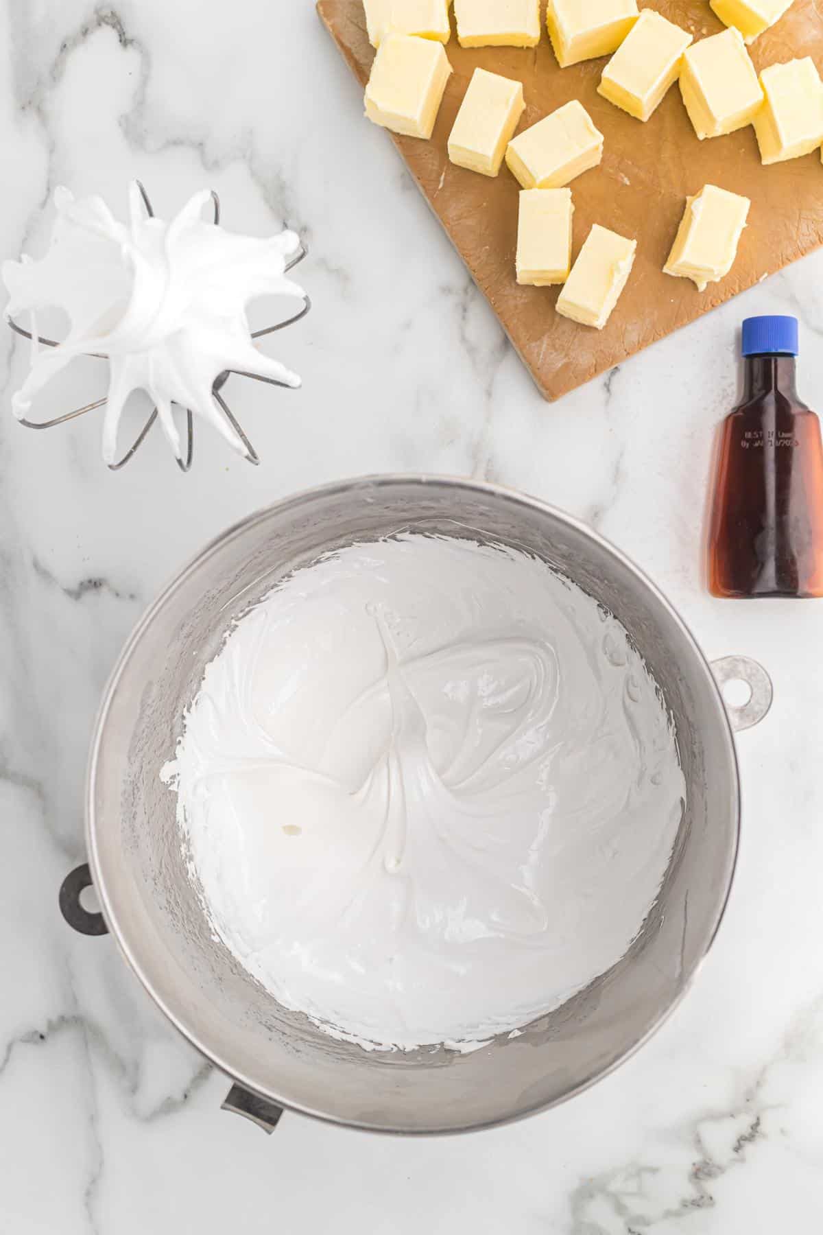 Mixer bowl filled with glossy white meringue with cubed butter and vanilla extract nearby
