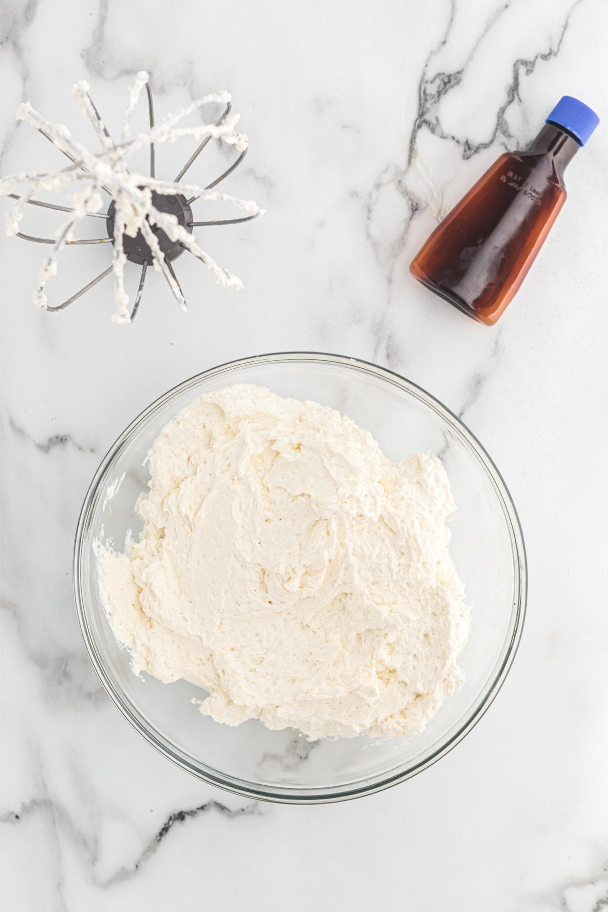 Glass bowl of finished buttercream frosting with a whisk attachment resting beside it