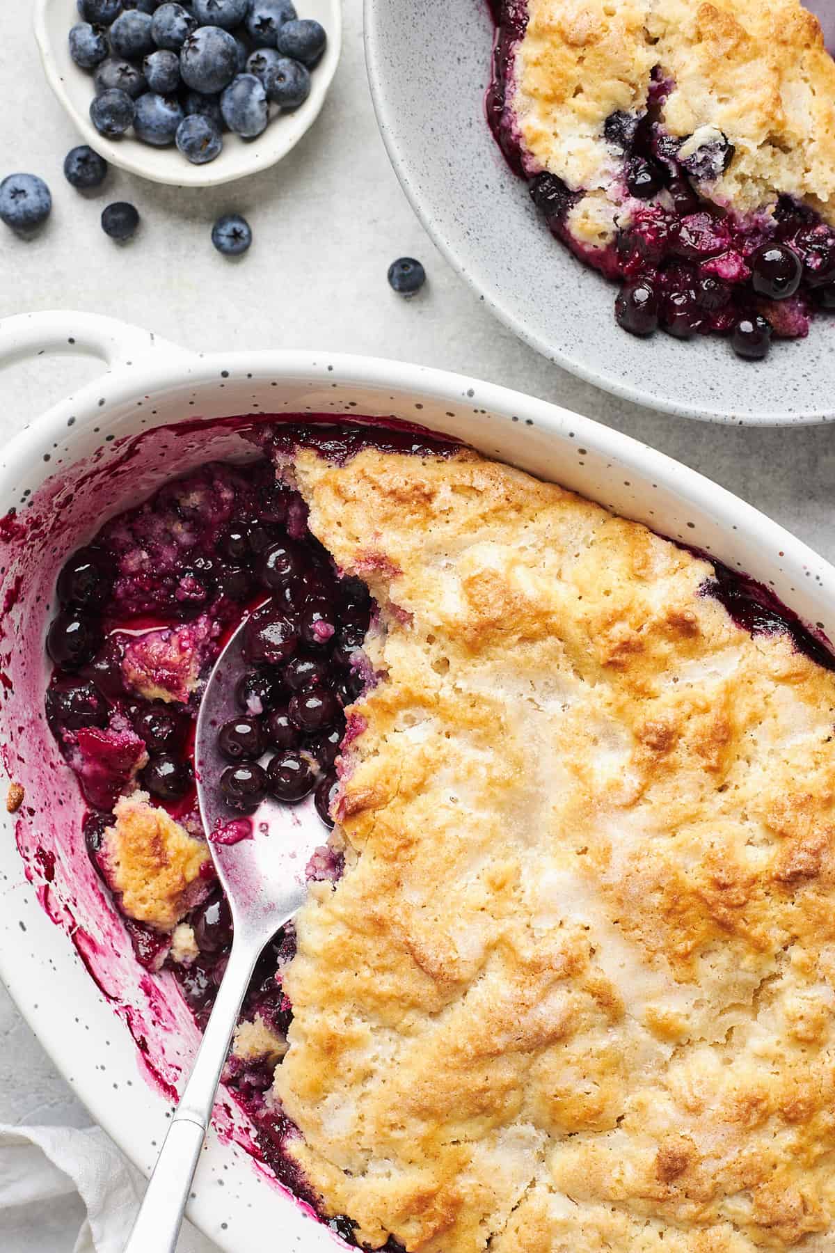 A serving spoon in a baking dish of fresh blueberry cobbler.
