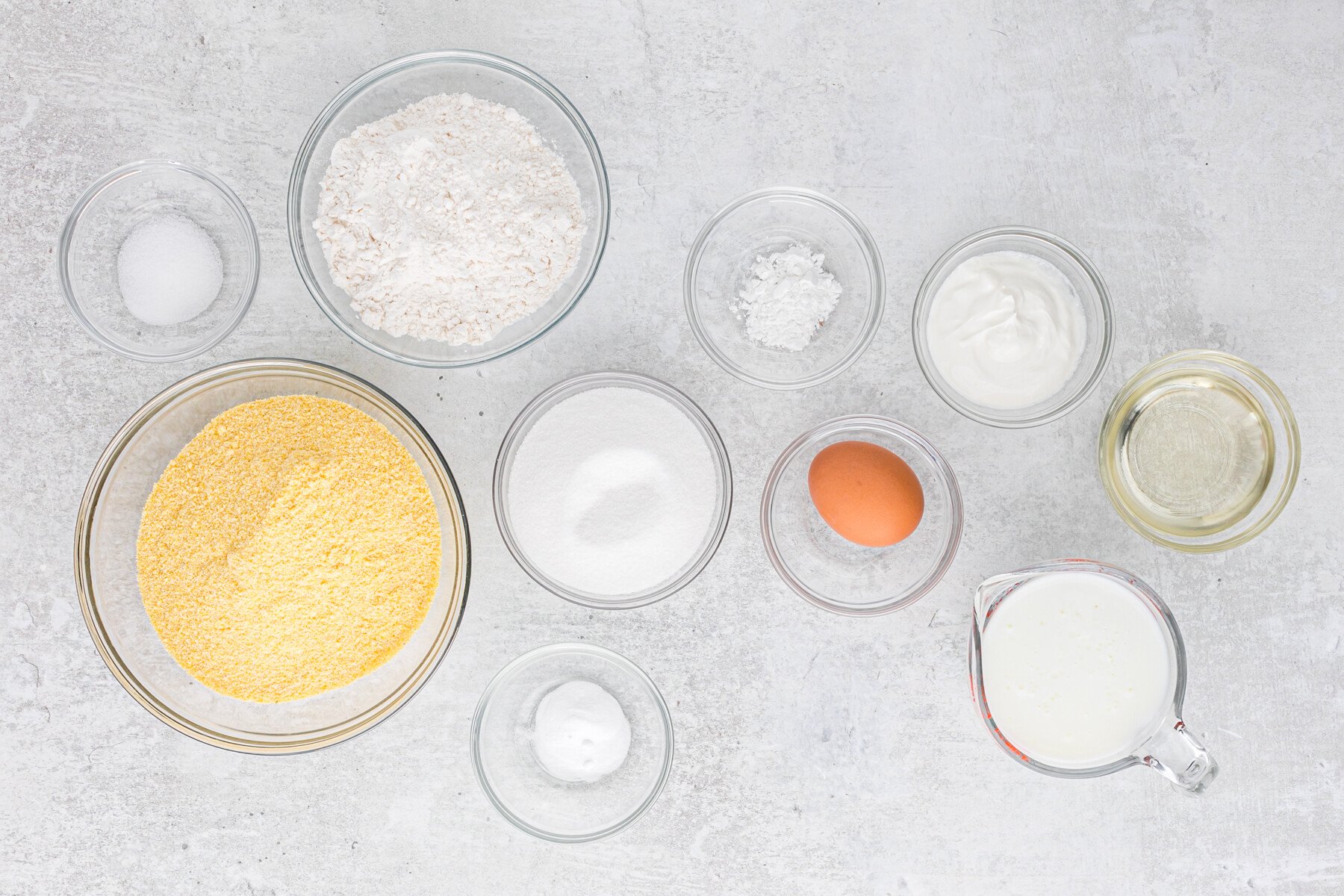 Cornmeal, flour, sugar, egg, leavening, oil and buttermilk in glass bowls on a gray background