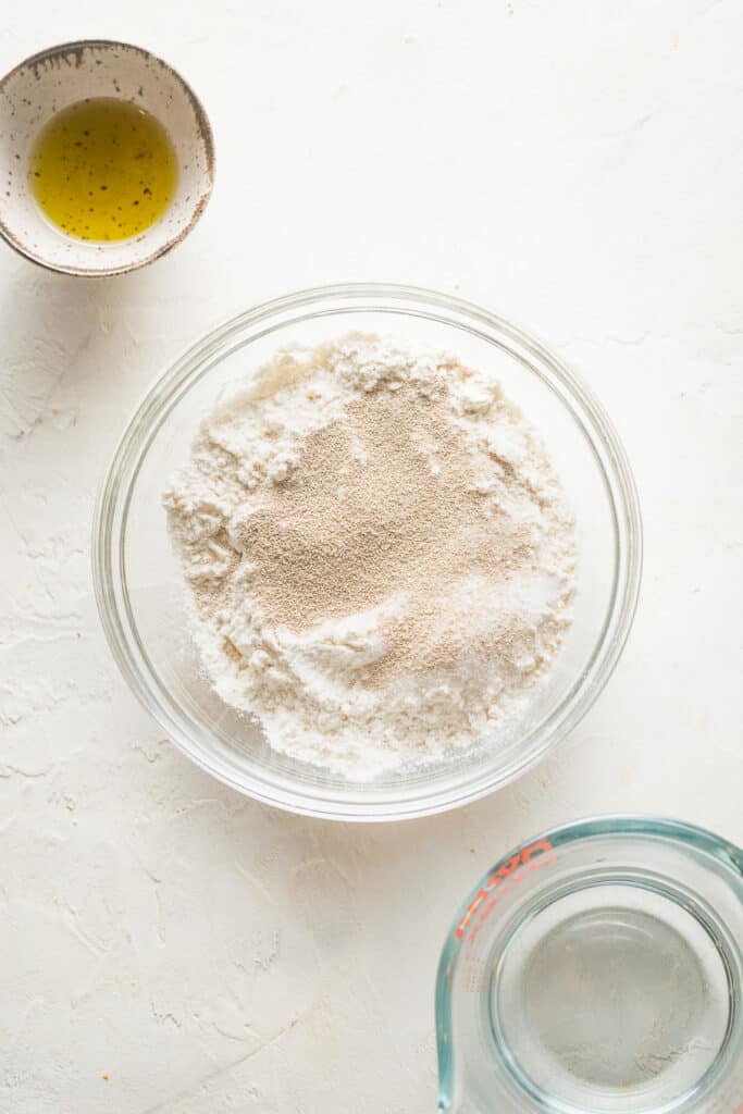 A glass bowl containing flour with a sprinkle of yeast on top, accompanied by a small bowl of olive oil and a measuring cup of water on a white background