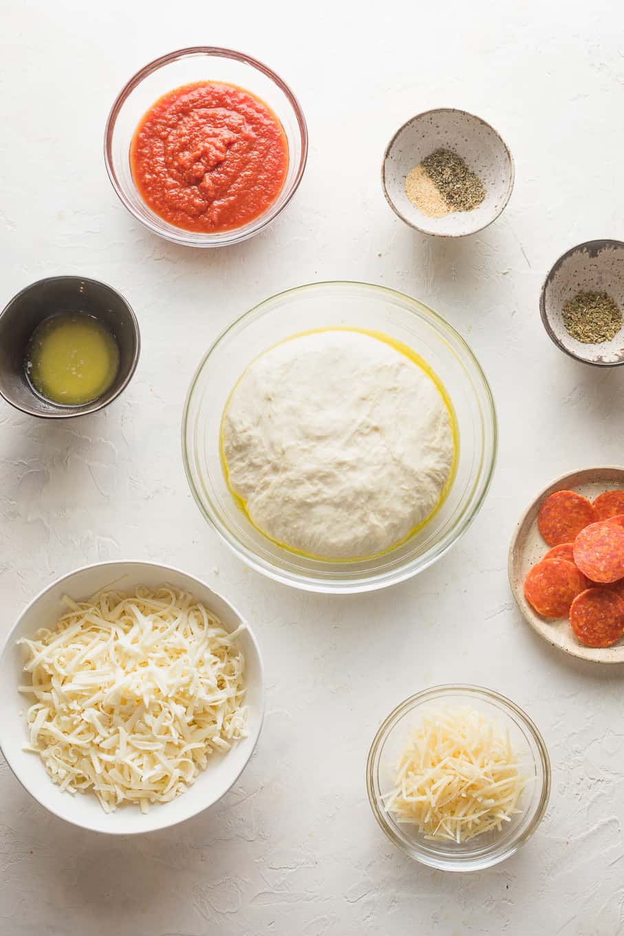 Ingredients for pepperoni pizza and crust in bowls on a white surface.