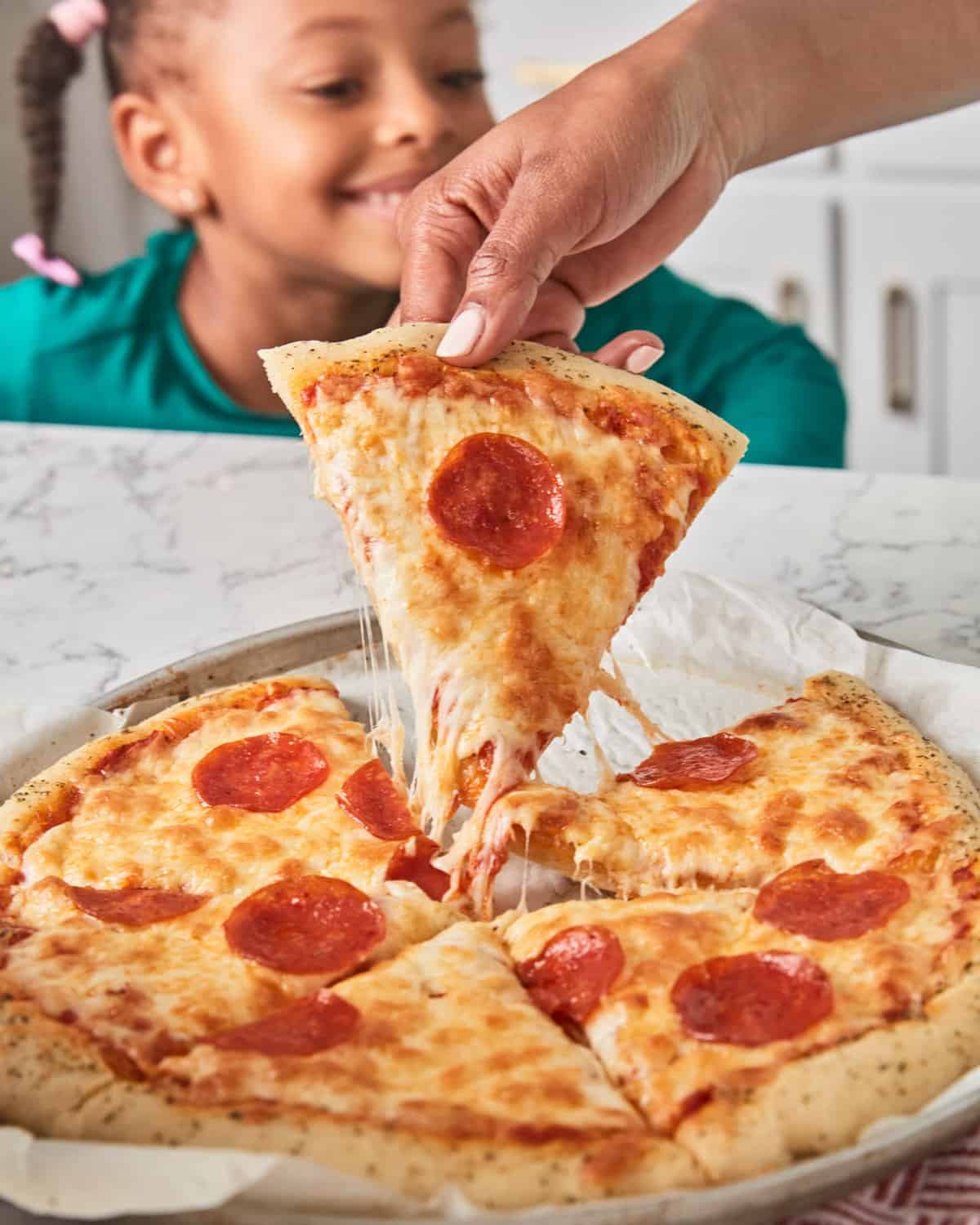 A closeup of a slice of pepperoni pizza with melted cheese stretching as it is lifted from the pizza, with a young girl smiling in the background