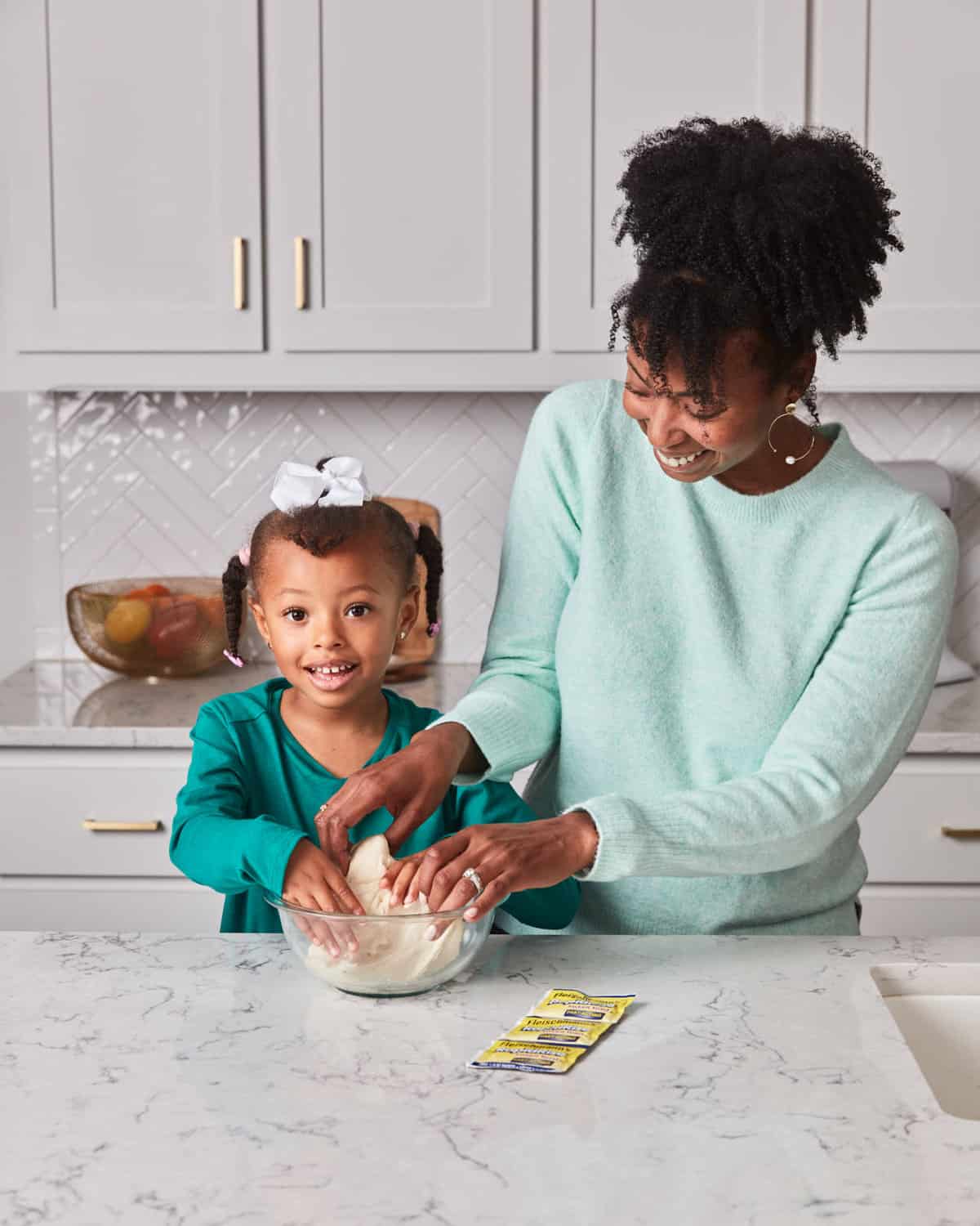 A black mom and her daughter smiling as they knead dough together in a bowl on a kitchen counter to make pepperoni pizza