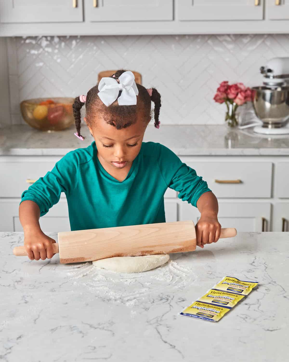 A young girl using a rolling pin on a pizza crust at the counter.
