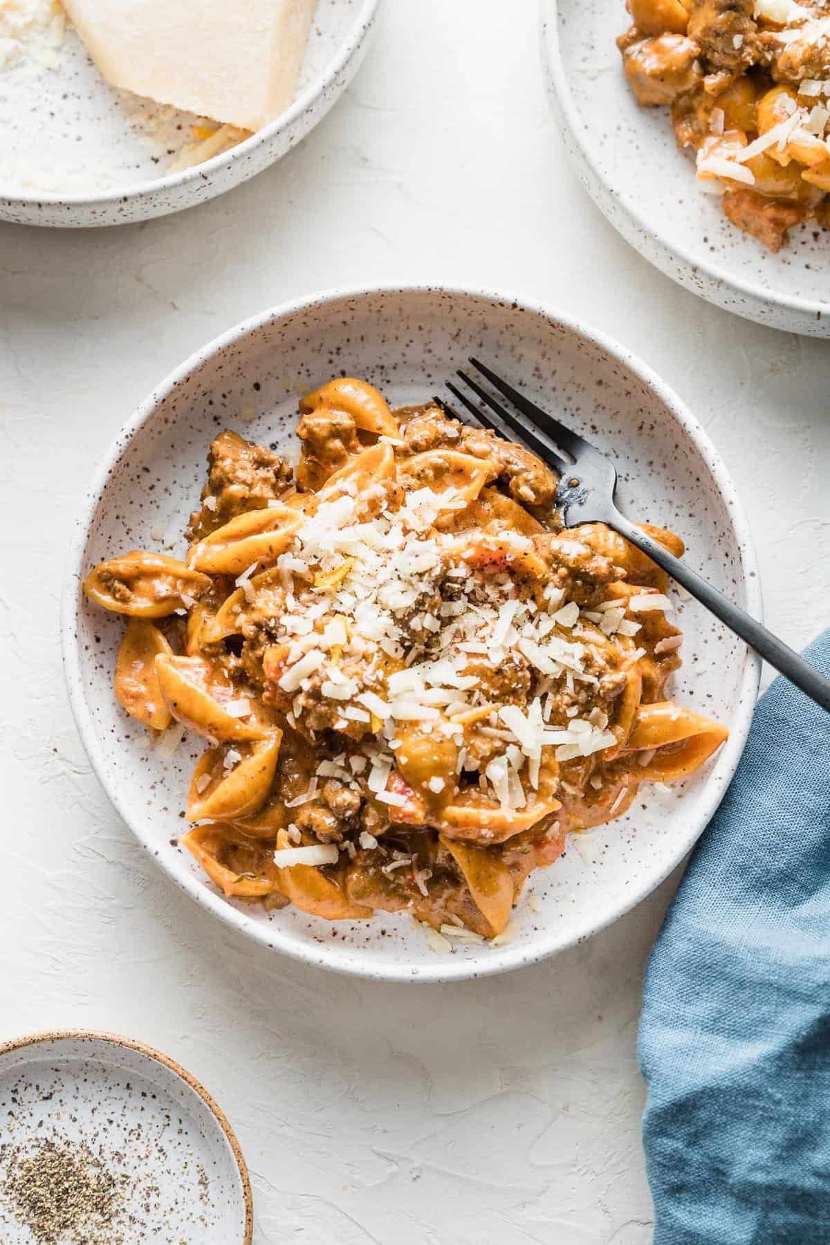 cheesy homemade hamburger helper recipe in a white speckled bowl with a fork on the side on a white background with a blue napkin.  