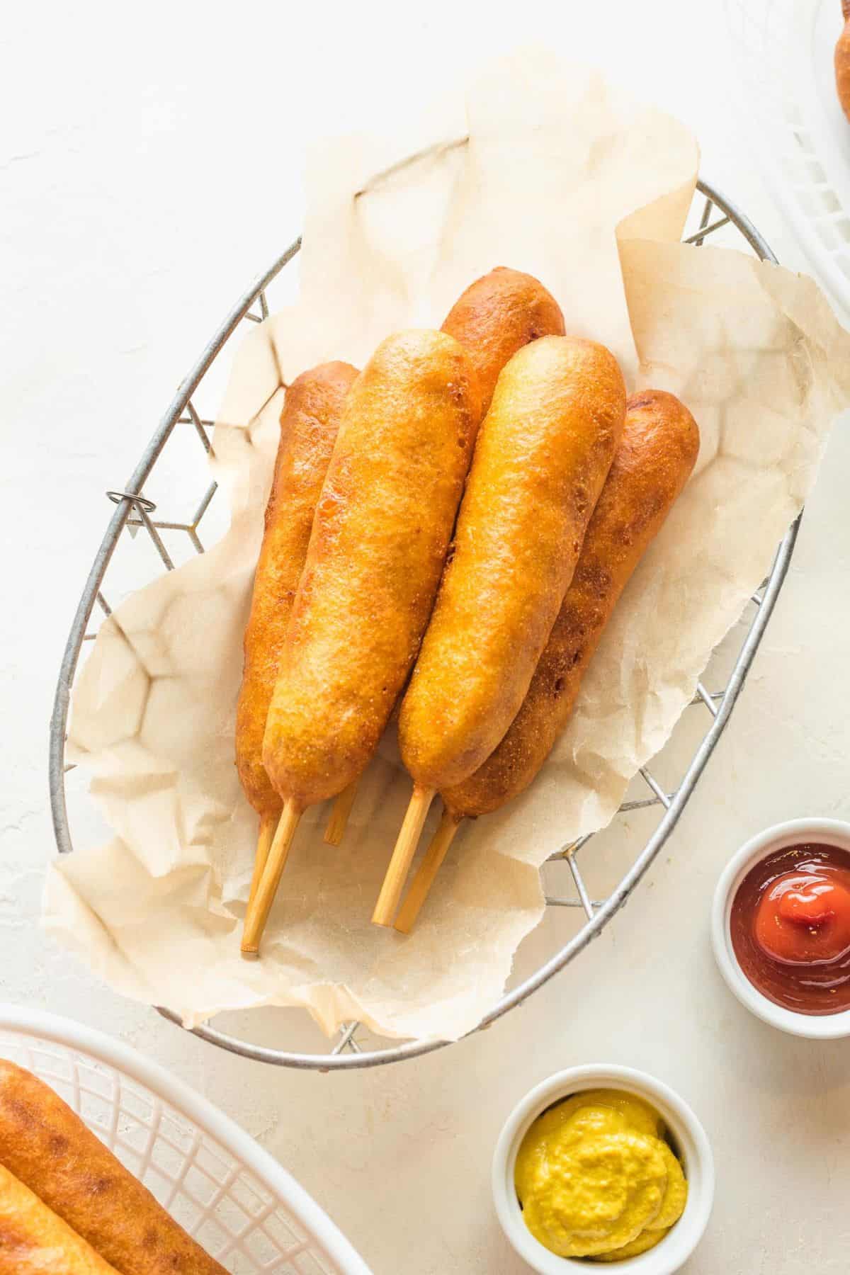 Golden-brown homemade corn dogs in a basket with small dishes of mustard and ketchup on the side