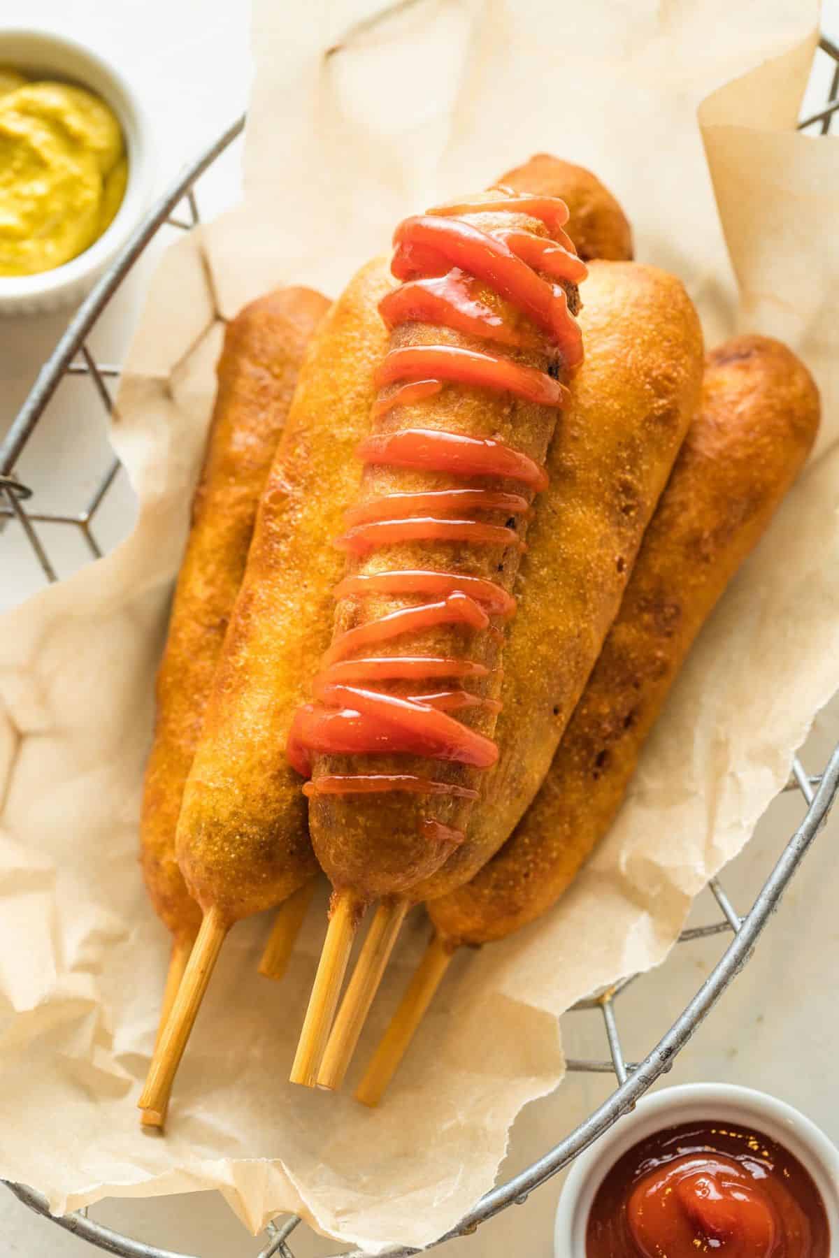 A homemade corn dog drizzled with ketchup on top of other corn dogs in a basket, with small dishes of mustard and ketchup on the side