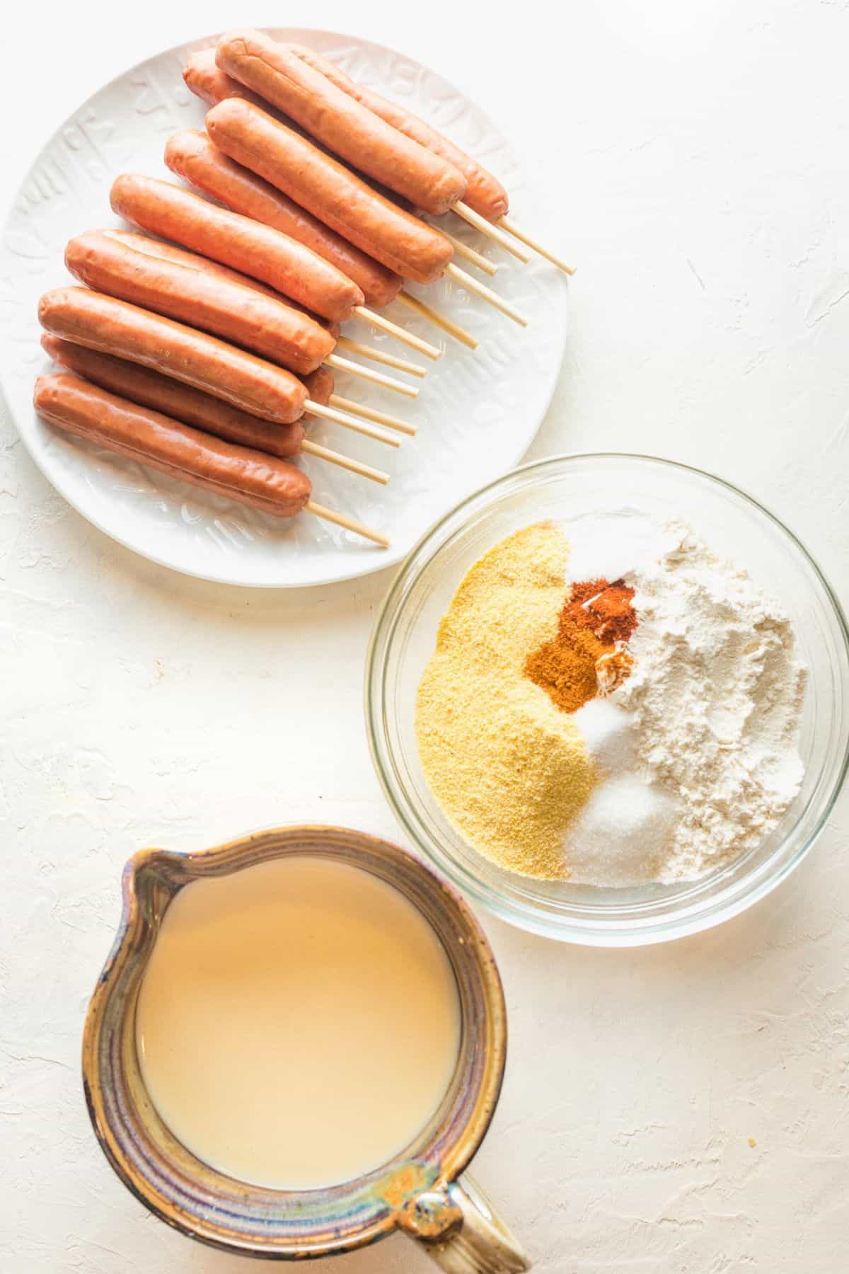 Overhead shot of ingredients to make homemade corn dogs on a white surface before cooking