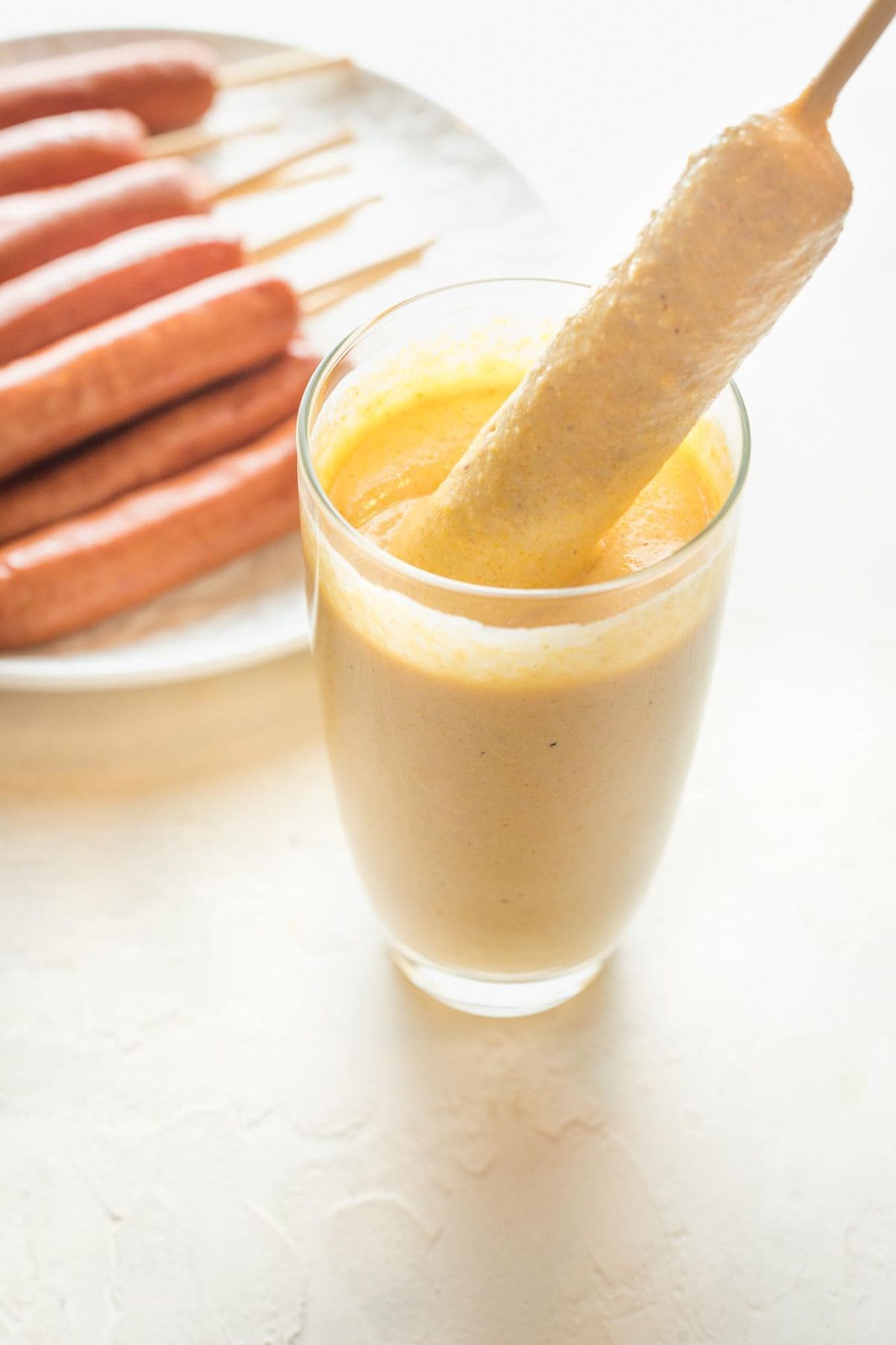 A skewered hot dog being dipped into a tall glass of batter, showing the batter-coating step