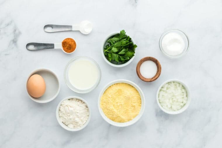 Cornmeal, flour, seasonings, parsley, onion, milk and egg in white bowls on white background