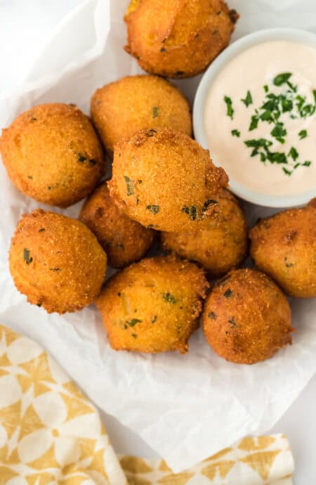 The best hush puppies fried on a paper towel with sauce in a white ramekin on a white background with yellow and white towel