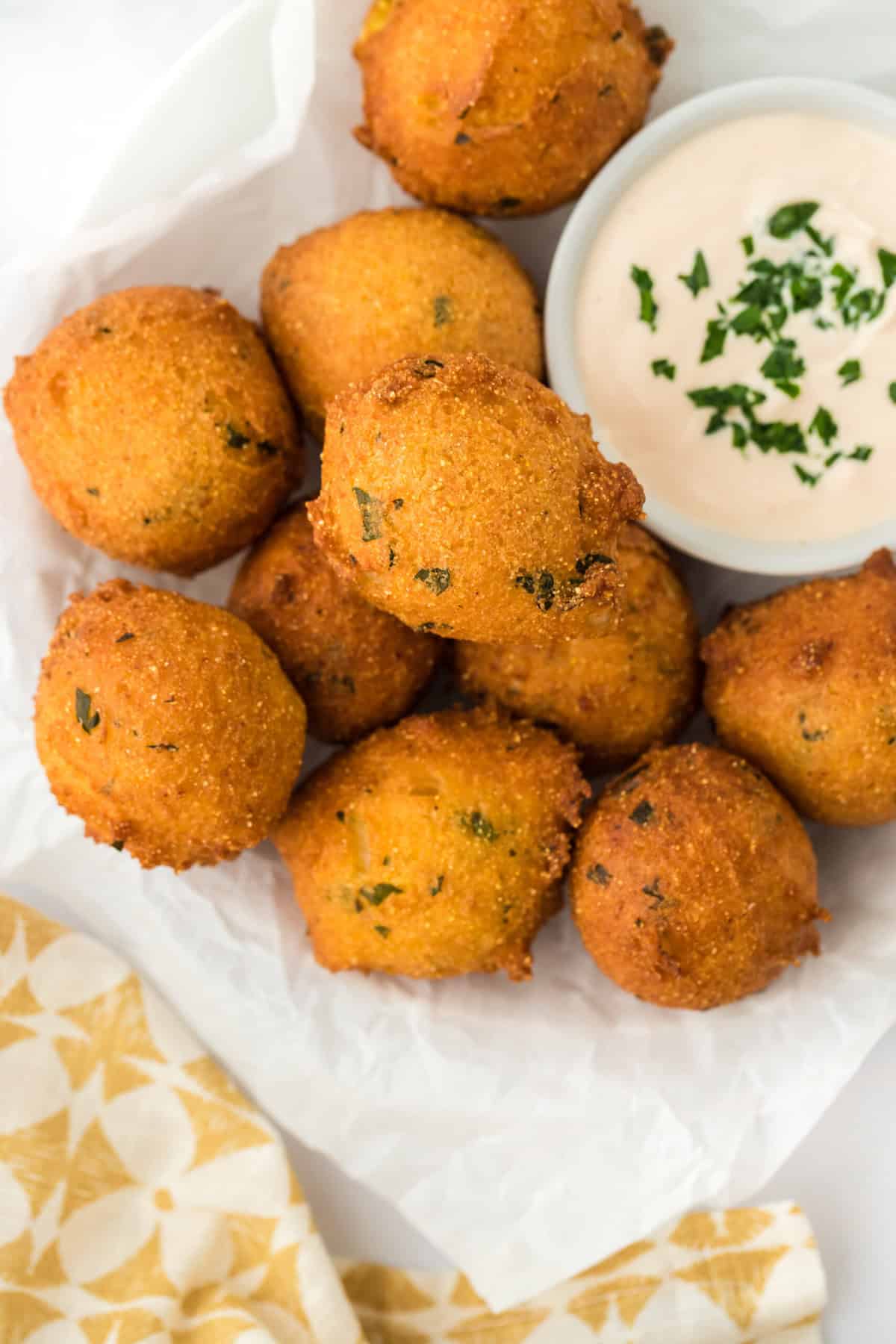 The best hush puppies fried on a paper towel with sauce in a white ramekin on a white background with yellow and white towel