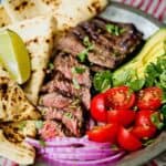 Sliced carne asada served on a platter with tortillas, tomatoes, avocado and onions.