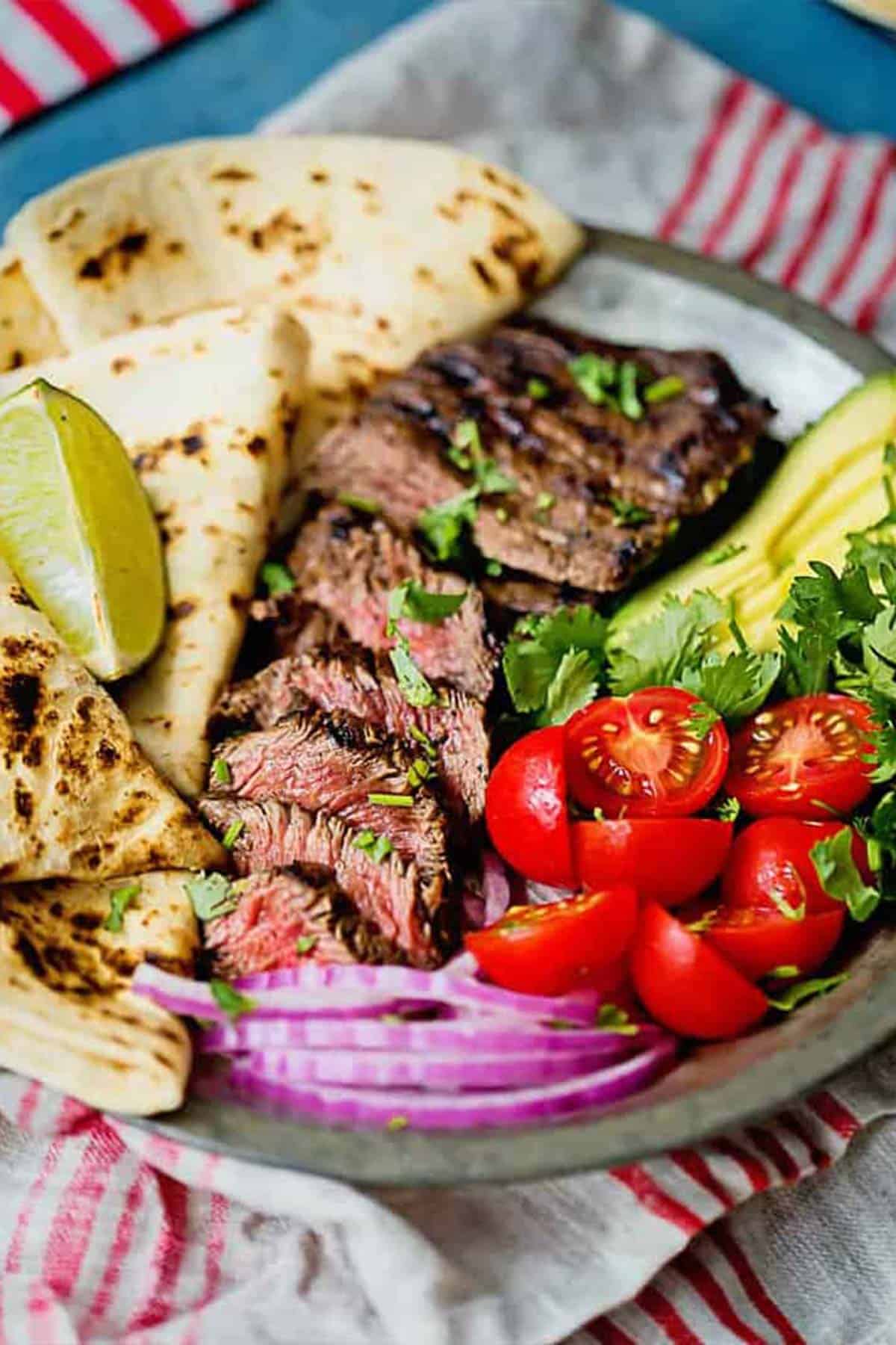 Sliced carne asada served on a platter with tortillas, tomatoes, avocado and onions.