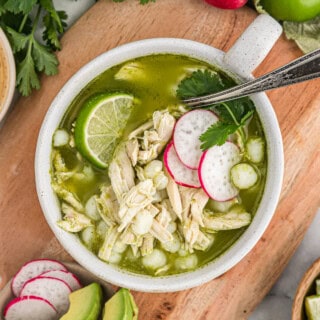 A small white bowl of chicken pozole verde on a wooden cutting board surrounded by avocado, limes and cilantro