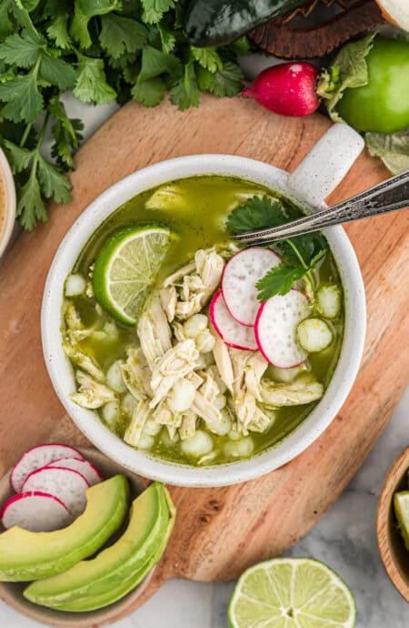 A small white bowl of chicken pozole verde on a wooden cutting board surrounded by avocado, limes and cilantro