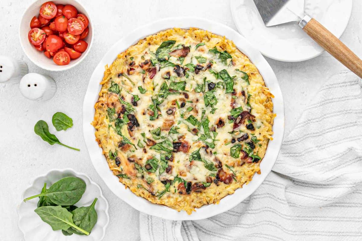 A bacon hash brown crust quiche in a white pie dish with a spatula on a plate and bowls of tomatoes and spinach on the side.
