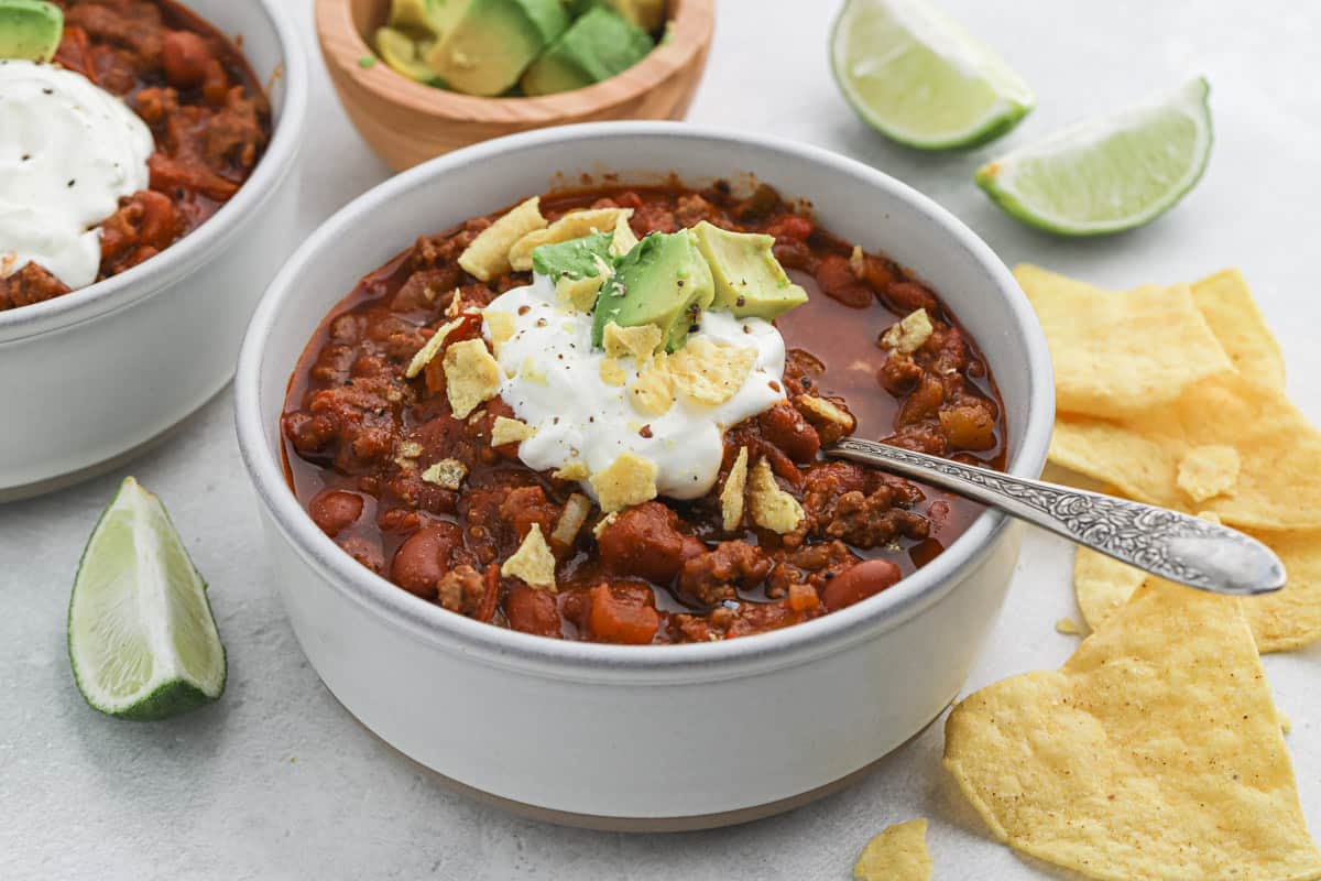 The best chili ever in two white bowls topped with sour cream, lime, avocado and tortilla chips ready to serve.