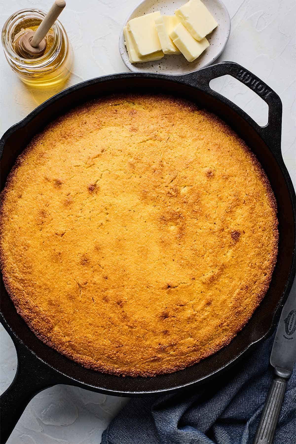 Homemade honey cornbread in a skillet on the table with a jar of honey and slices of butter on a small plate.