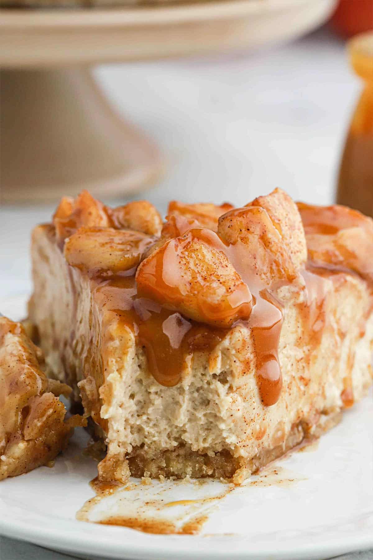 A decadent apple cheesecake with slice taken out on white plate with caramel sauce poured over it form the front.