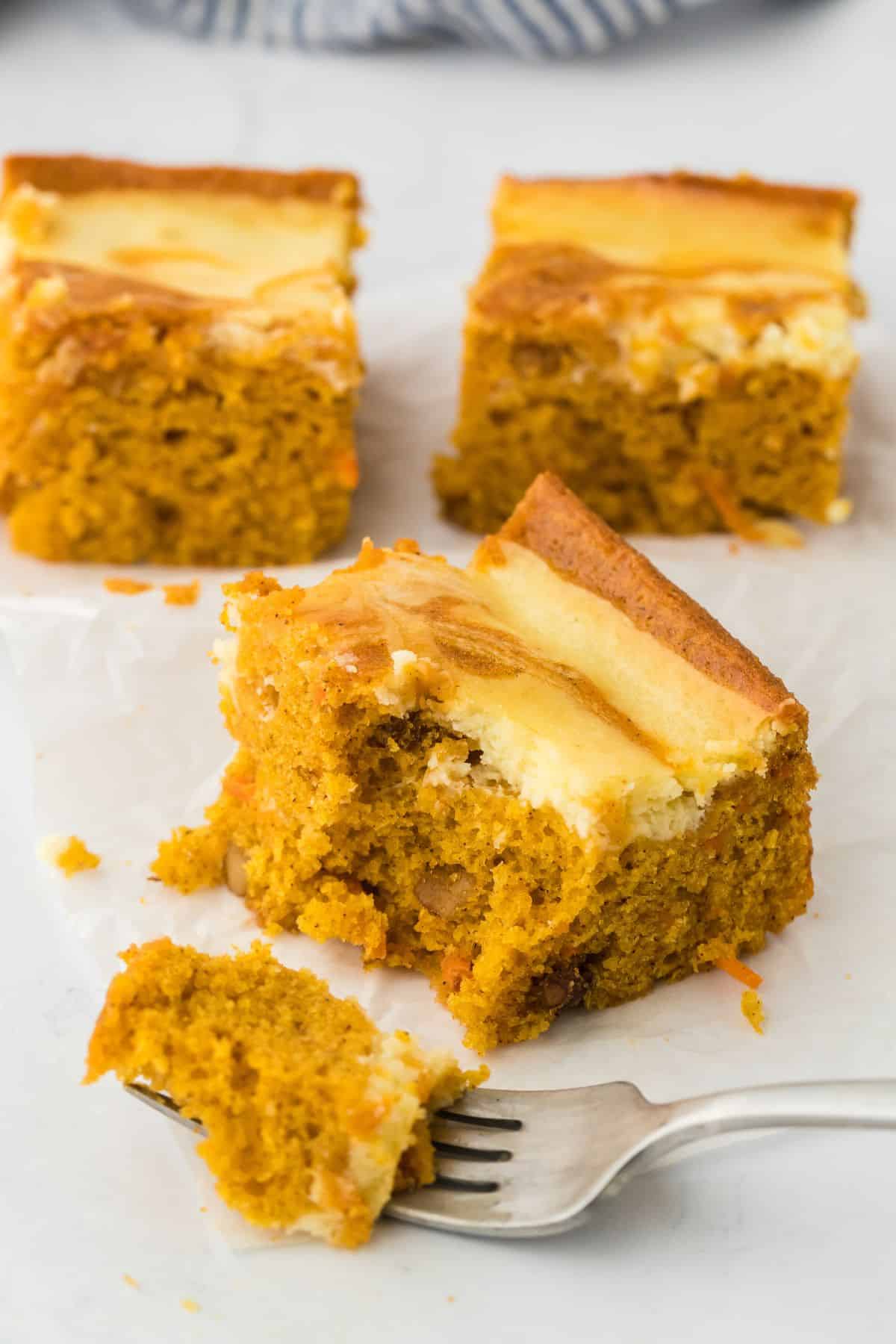 Three carrot cake swirl bars sitting on white parchment with a fork in the front with a bite on it. 