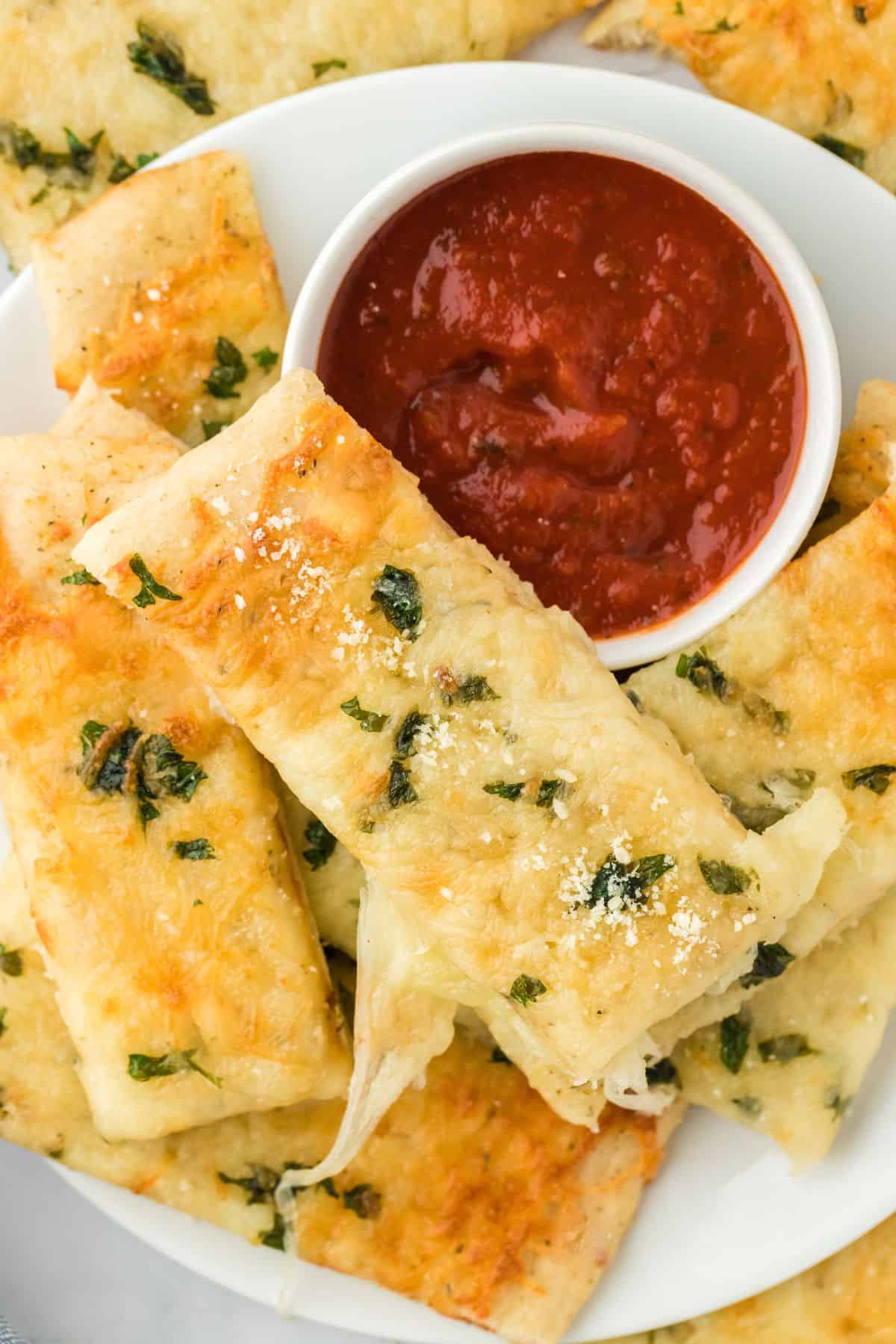A plate of cheesy garlic breadsticks on a plate with a small bowl of marinara sauce.