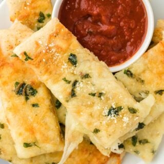 Cheesy garlic breadsticks on a plate with a bowl of pizza sauce.