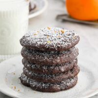 A stack of chocolate orange cookies on a plate in front of a glass of milk and an orange.