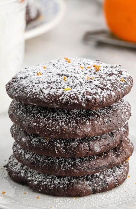 A stack of chocolate orange cookies on a plate in front of a glass of milk and an orange.