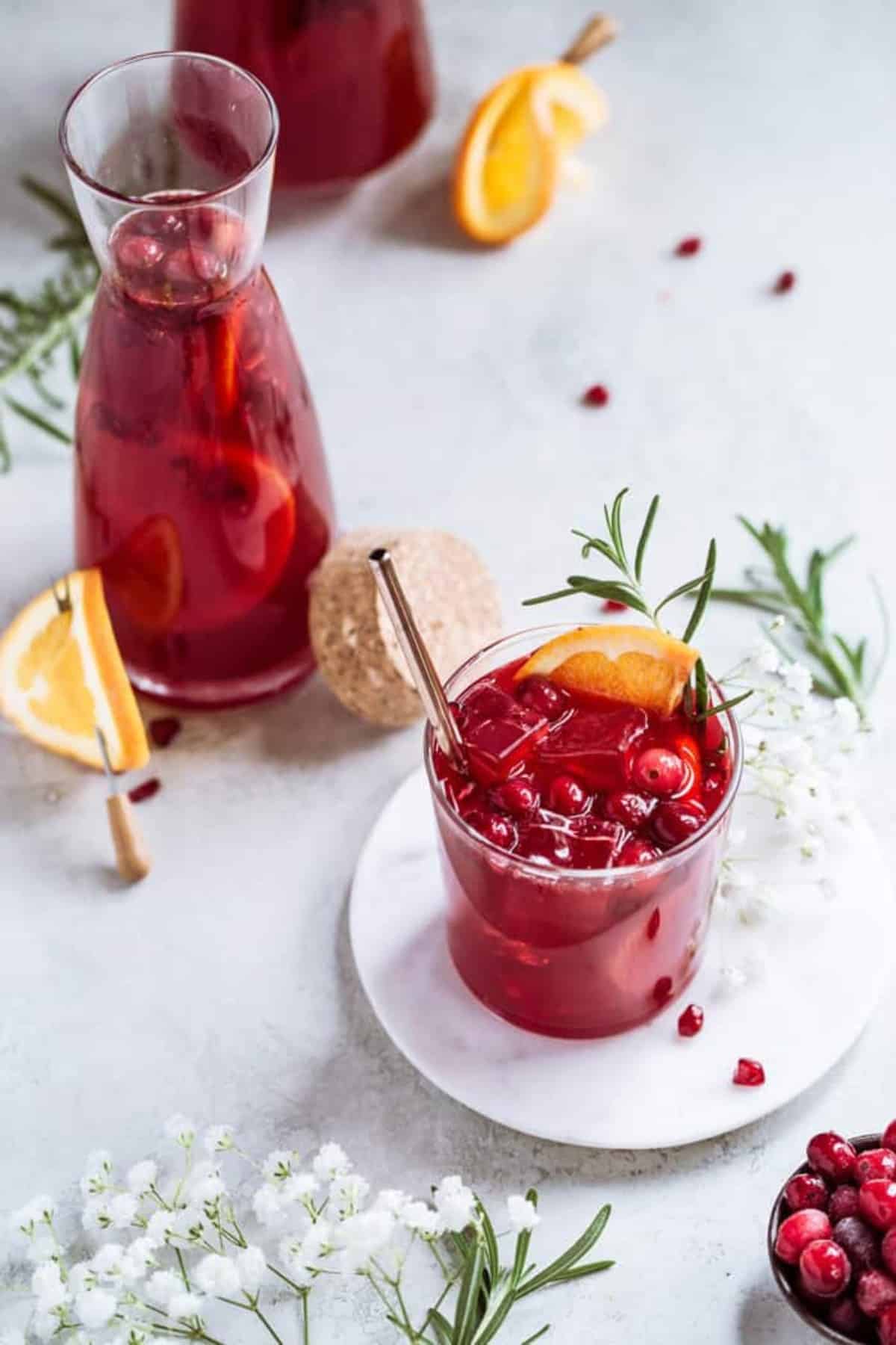 A glass of red pomegranate punch next to a filled carafe with rosemary nearby