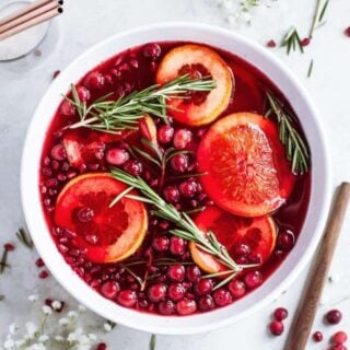 An overhead of a white punch bowl filled with Christmas punch and garnished with cranberries, pomegranate seeds, orange slices and rosemary