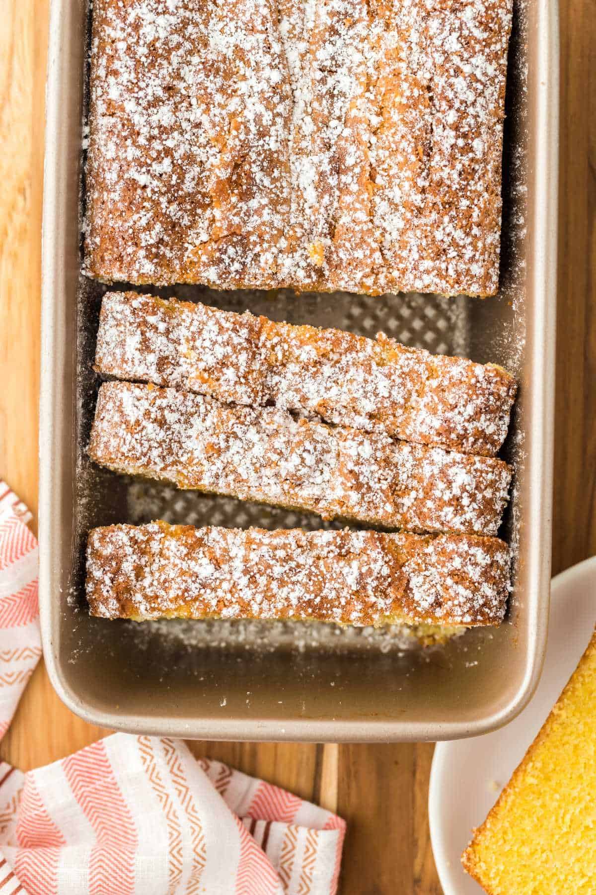 A loaf of cornmeal cake in a pan sprinkled with powdered sugar and a few slices cut and leaning forward in the pan.