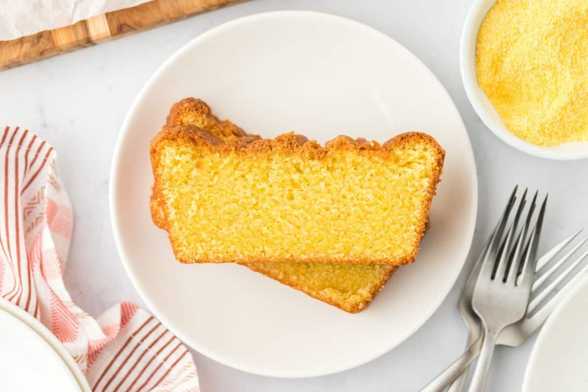 Two slices of cornmeal cake stacked on a plate with forks on the side and a small bowl of cornmeal.