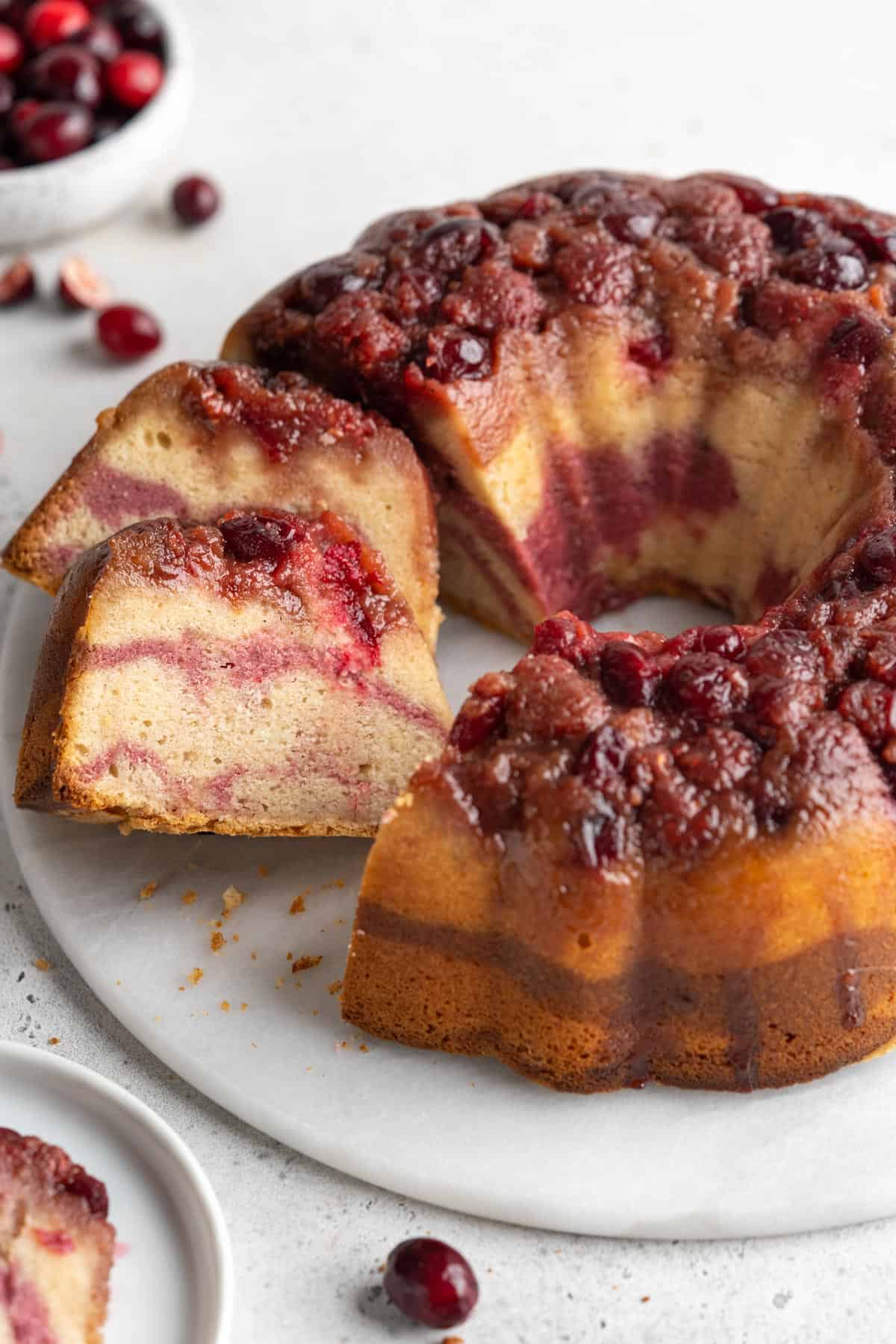 The cranberry bundt cake with slices cut out of it and leaning over on the platter.