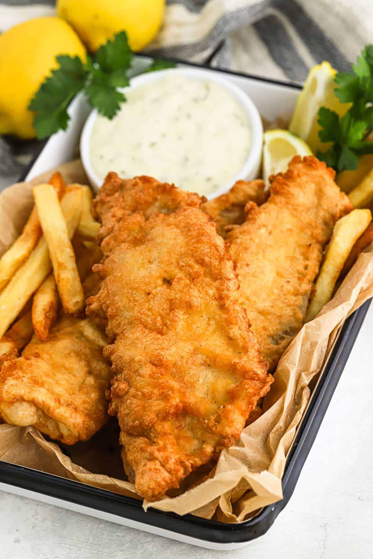 A tray with a portion of fish and chips with tartar sauce in the back.