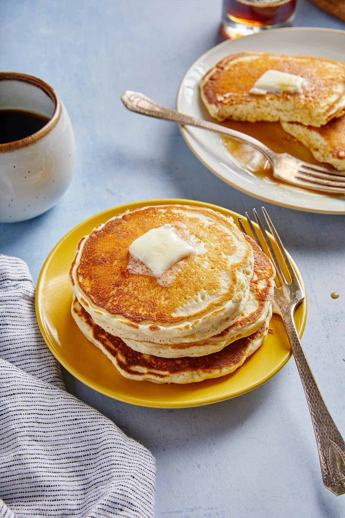 An overhead photo of a stack of classic pancakes with a melting pat of butter on top and a plate in the background partially eaten.